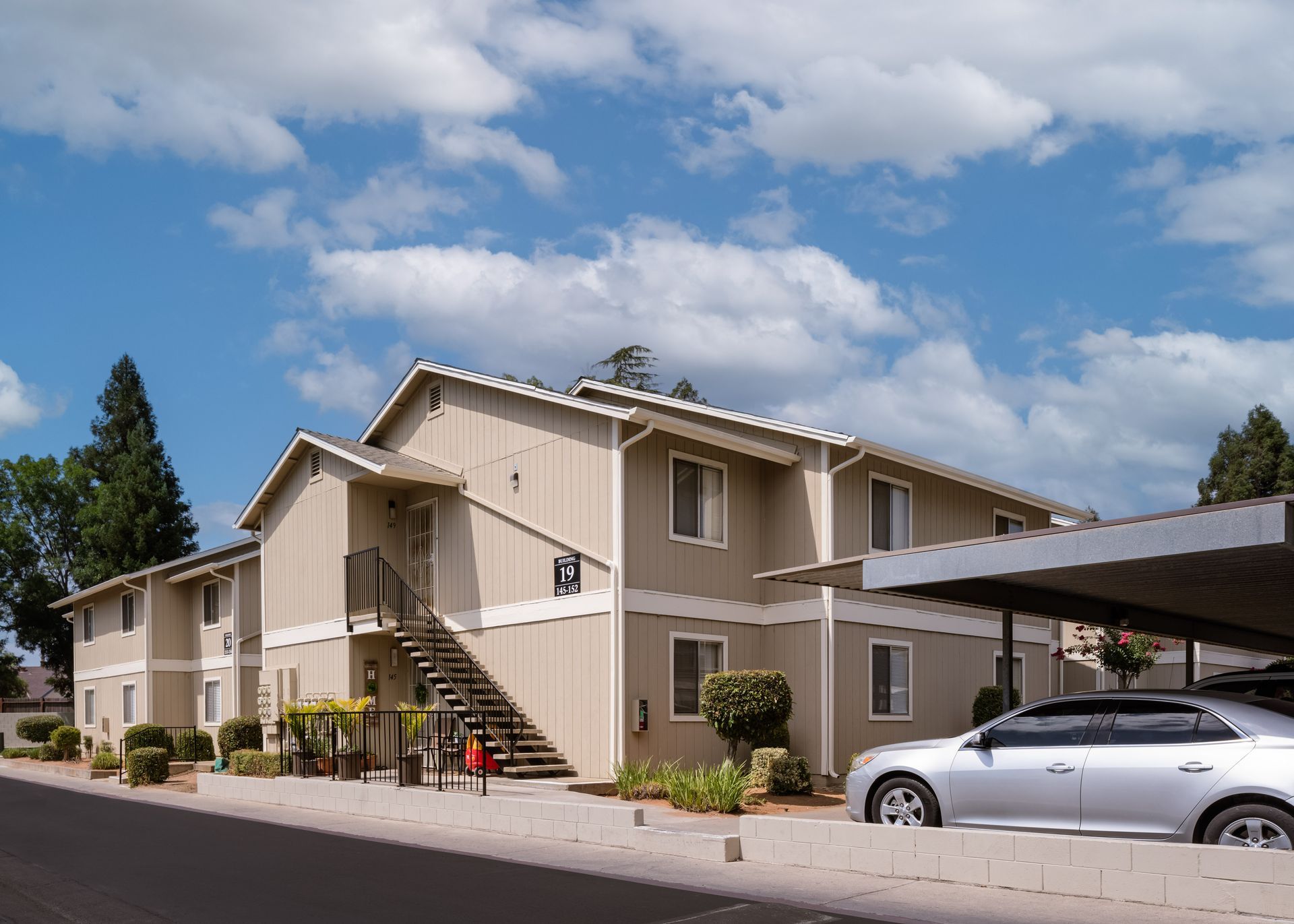 Apartment building with beige siding, car under a carport, and blue sky with clouds.