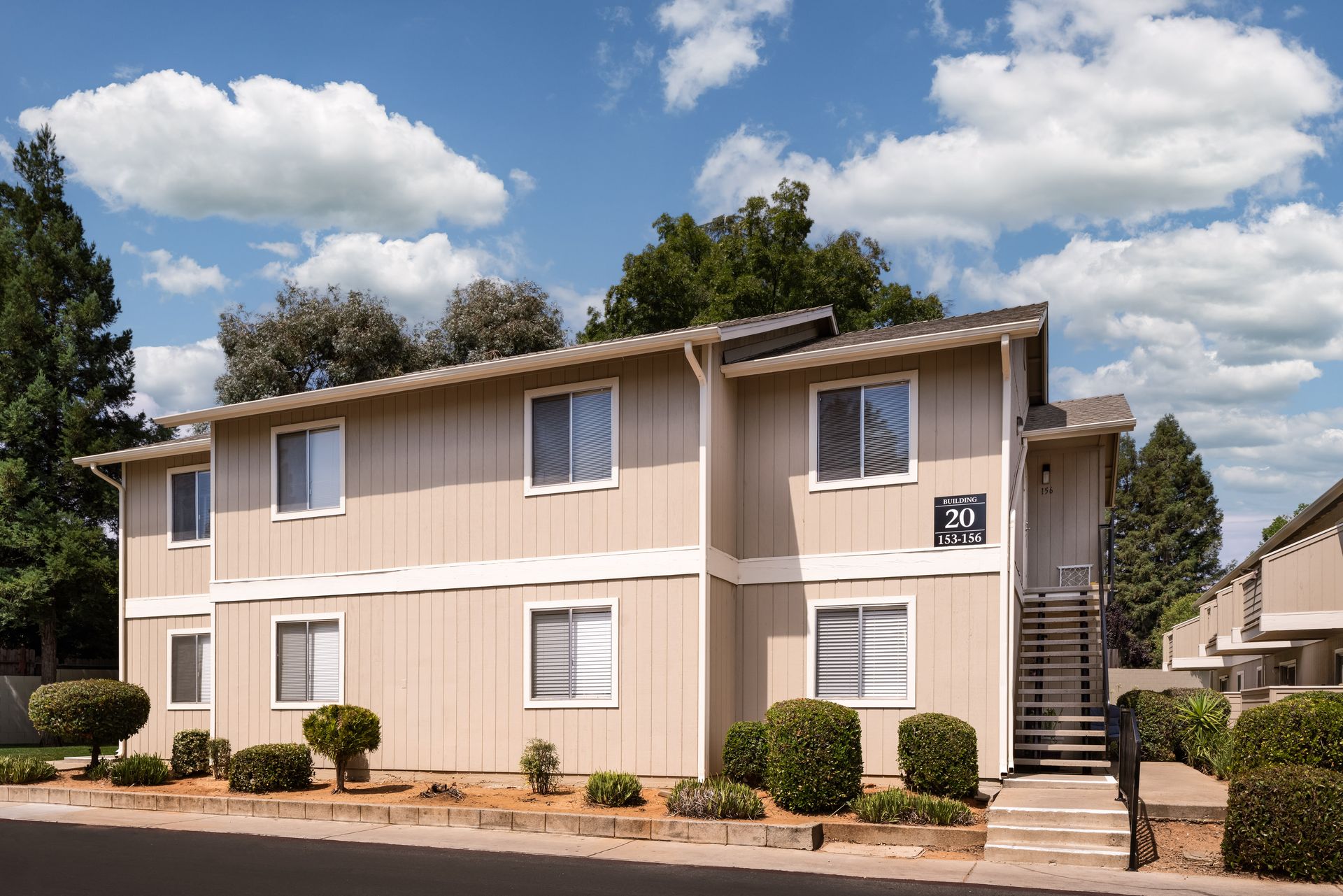 Two-story beige apartment building with windows and shrubbery under a blue sky.