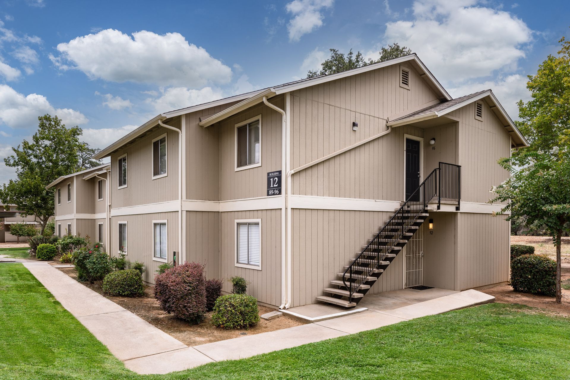 Two-story tan apartment building with outdoor stairwell and green lawn under a cloudy sky.