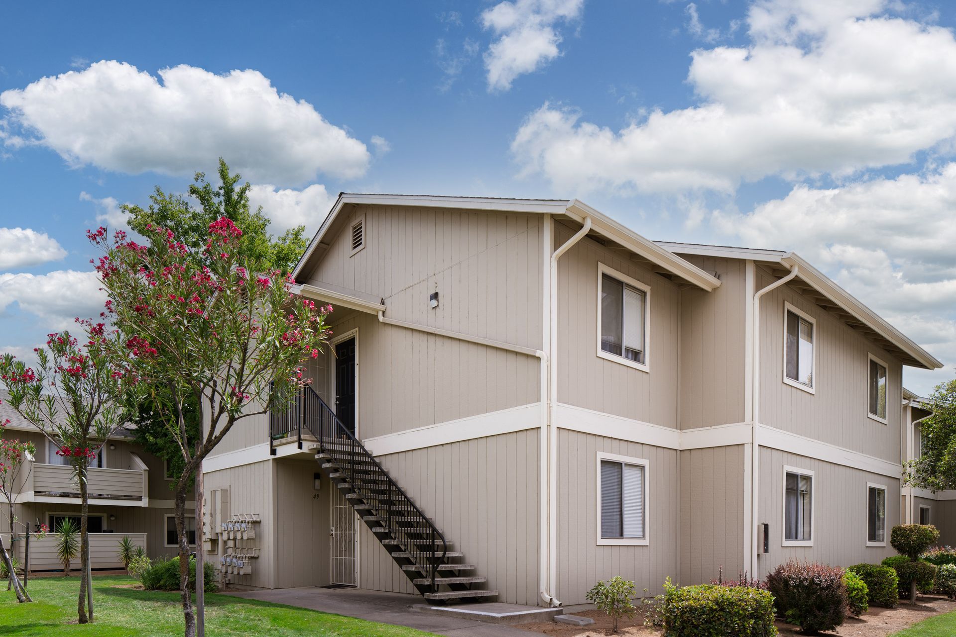 Two-story beige apartment building with black stairs, green lawn, and a blue sky with clouds.