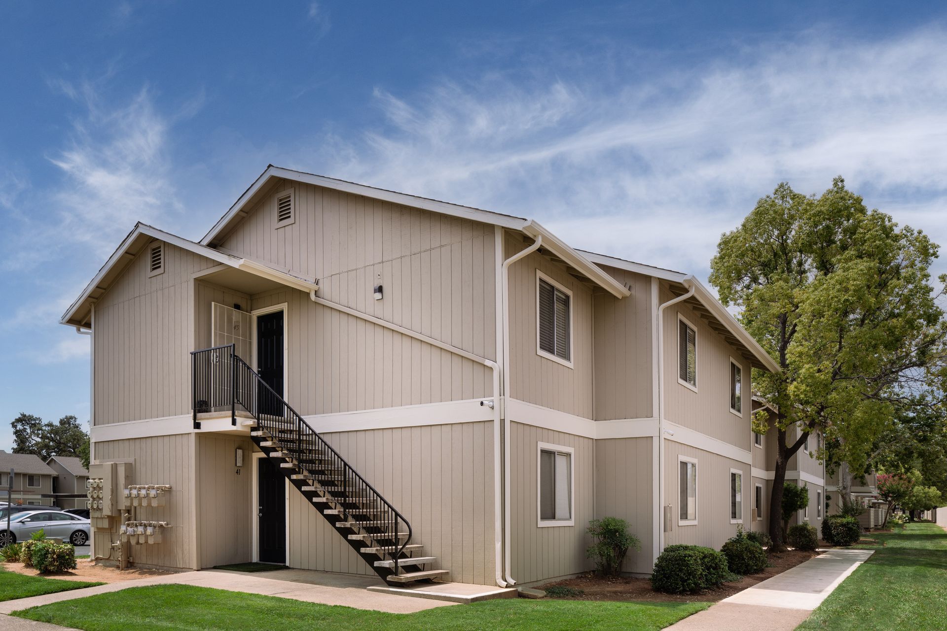 Two-story beige apartment building with exterior staircase, blue sky, and green lawn.