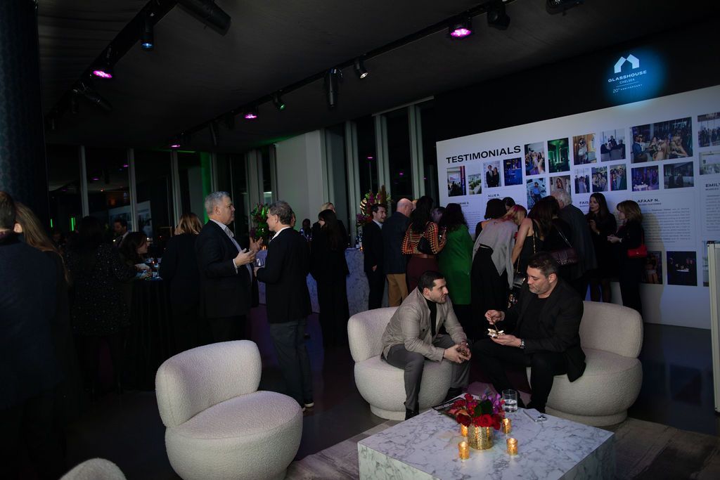 People at an event. Some are seated on white, rounded chairs. A wall with photos is visible.