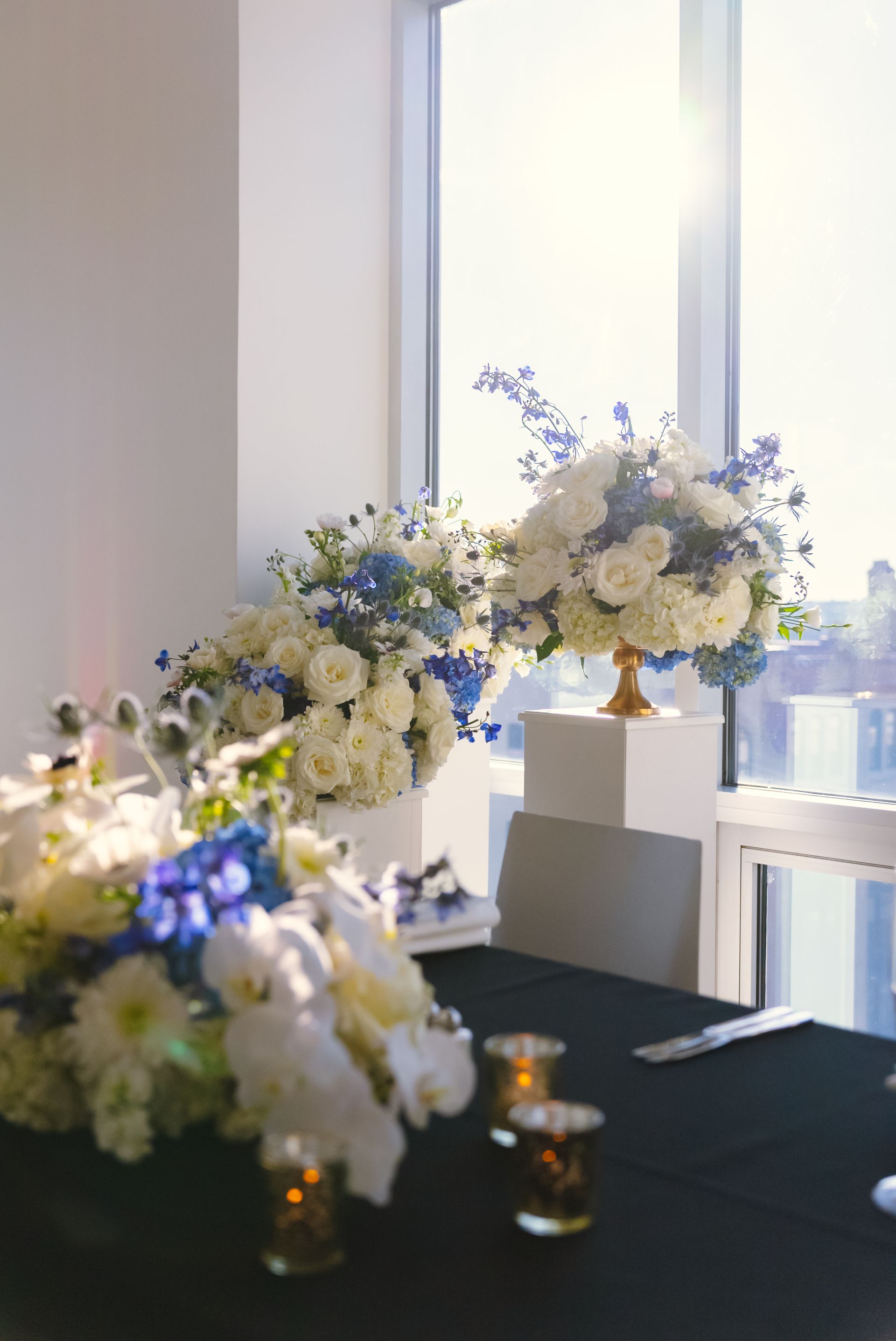 Elegant table setting with white and blue floral arrangements; tall window with sunlight.