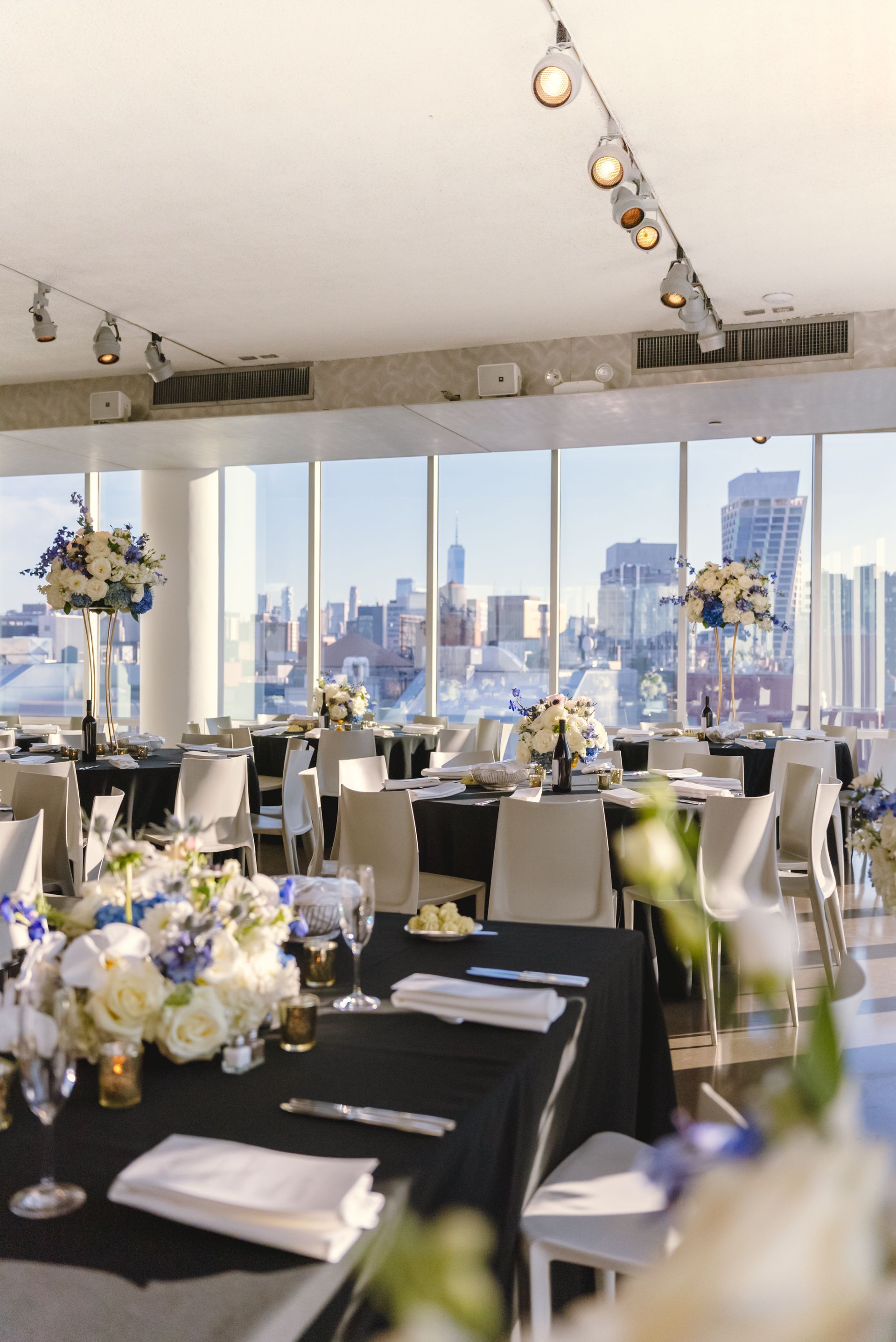 Wedding reception with tables set for guests, city skyline visible through windows. Blue and white floral centerpieces.