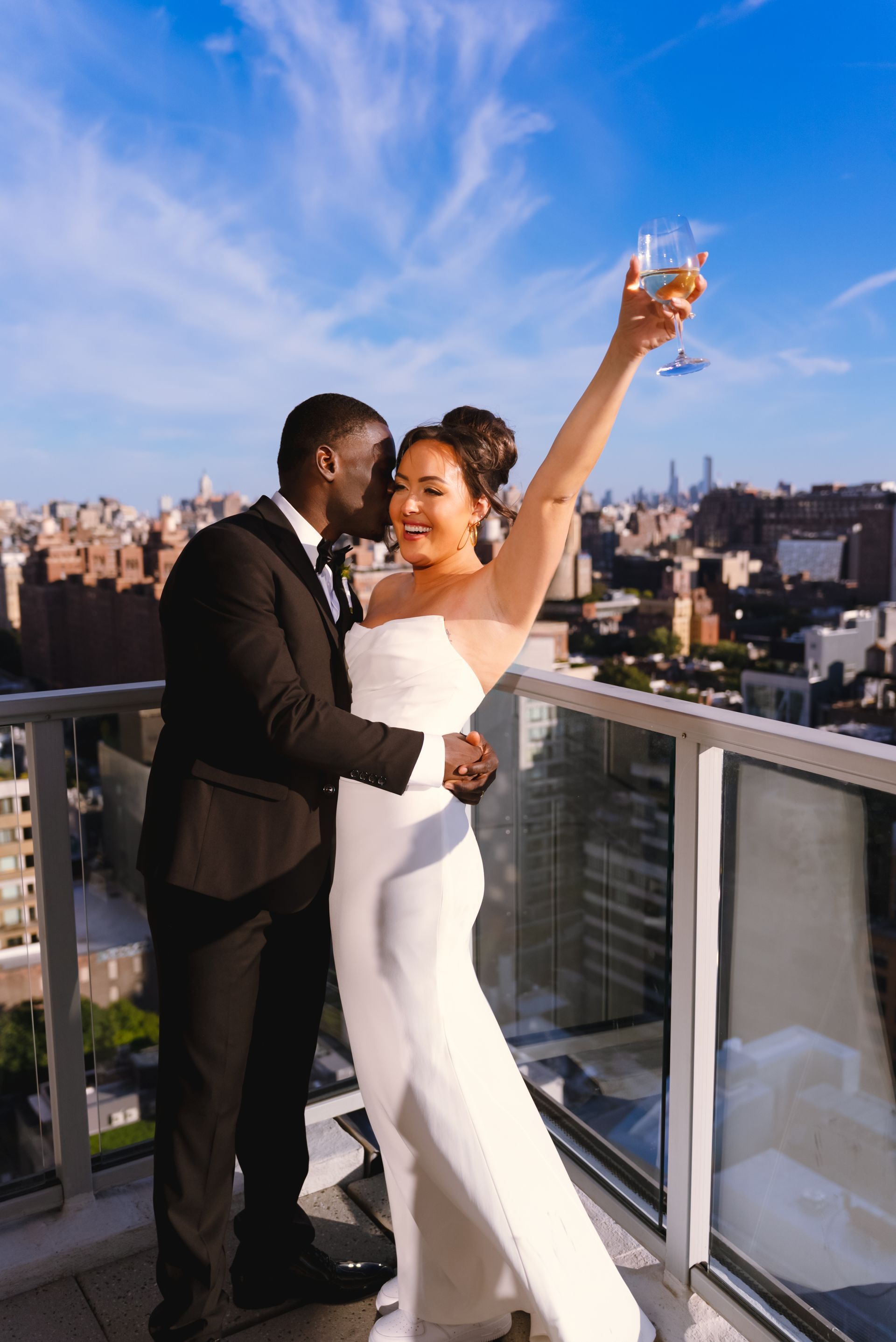 Couple on balcony celebrating, bride in white dress raises glass. City skyline background.
