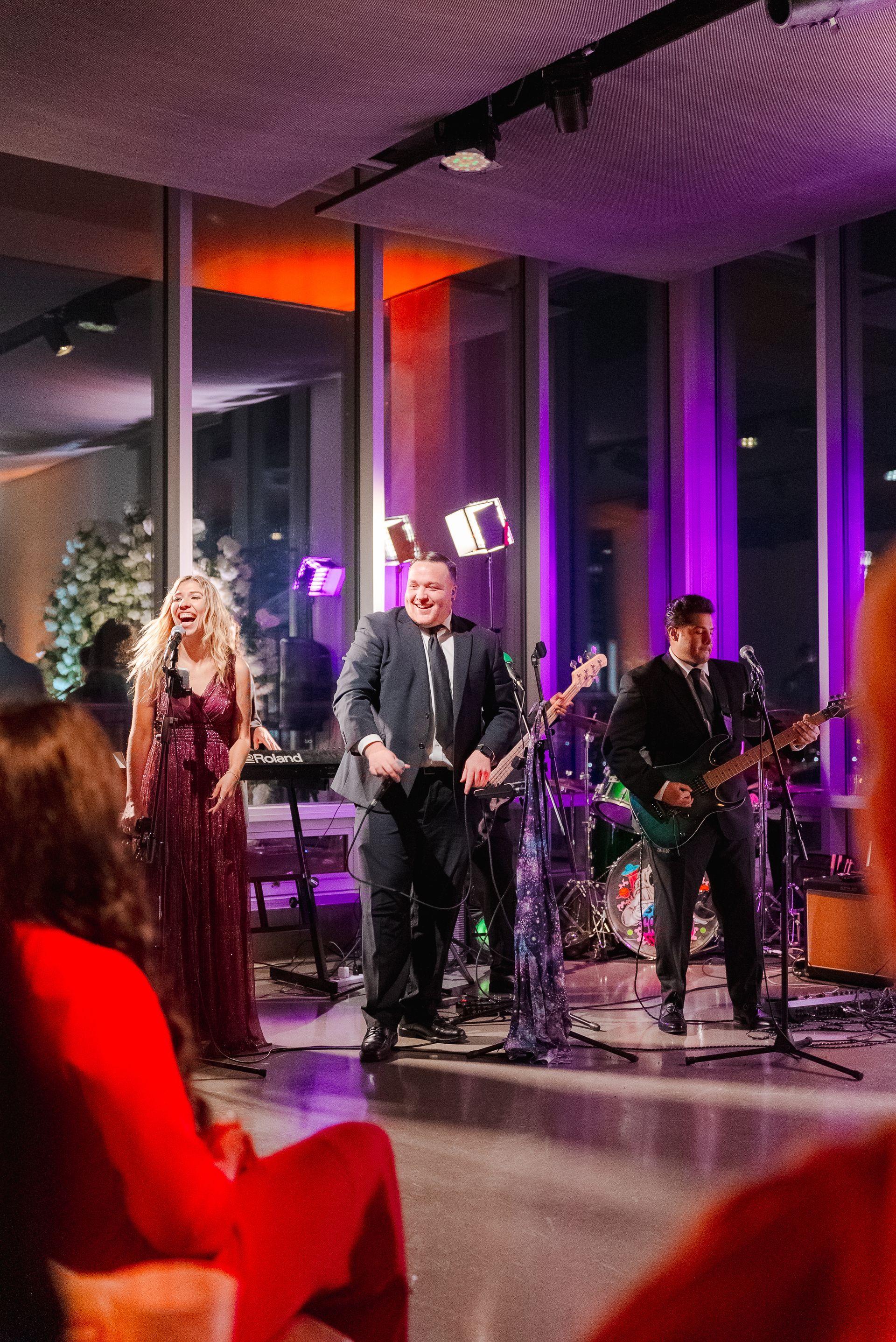 Band performing on stage in a room with large windows, featuring a singer, keyboardist, guitarist, drummer, and a singer.