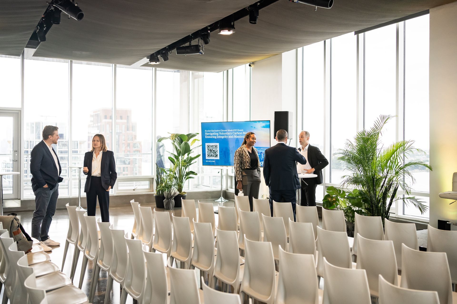 Conference room with rows of chairs, people talking near a screen and plants.