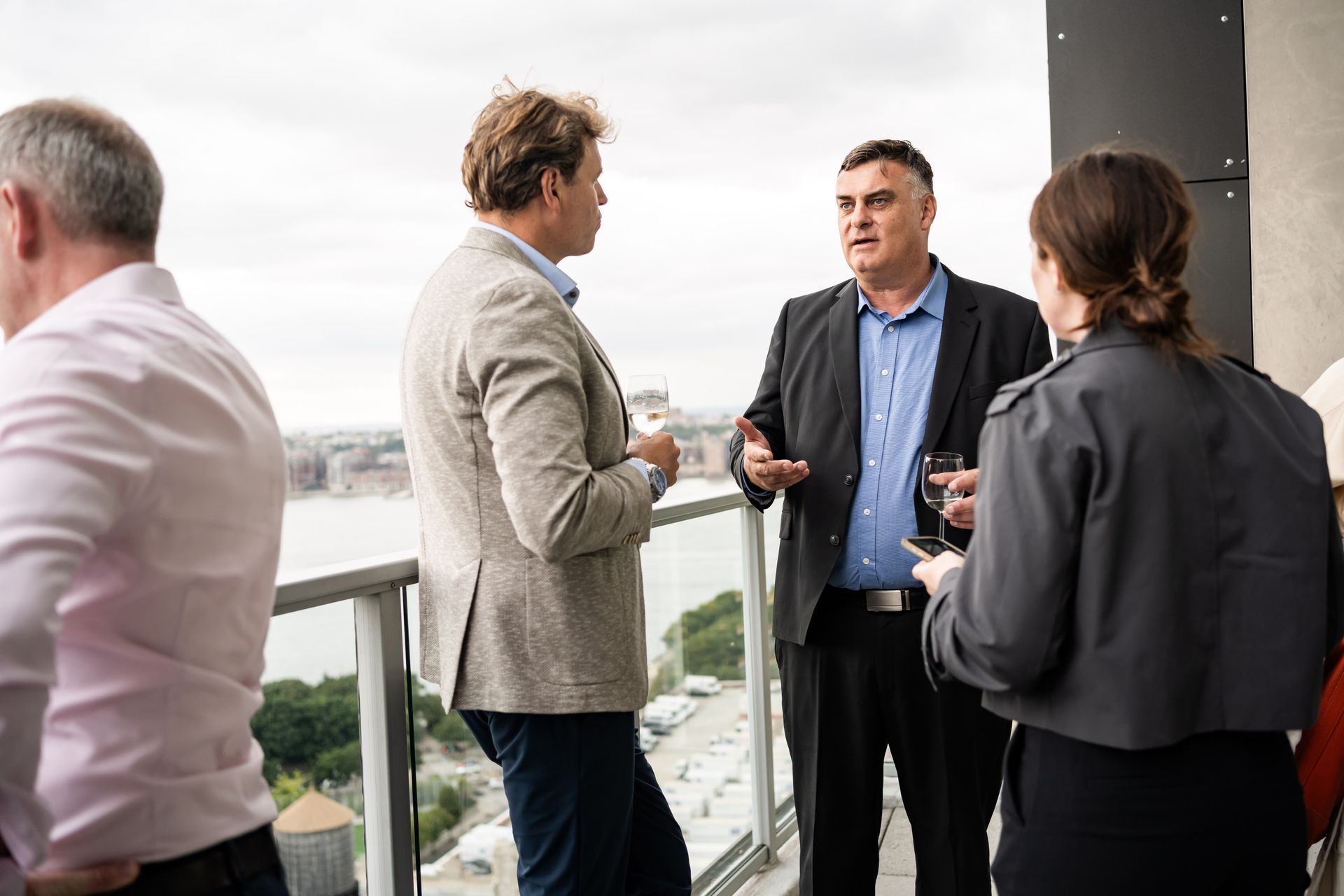 People conversing on a balcony overlooking a cityscape. A man gestures while speaking. Cloudy sky.