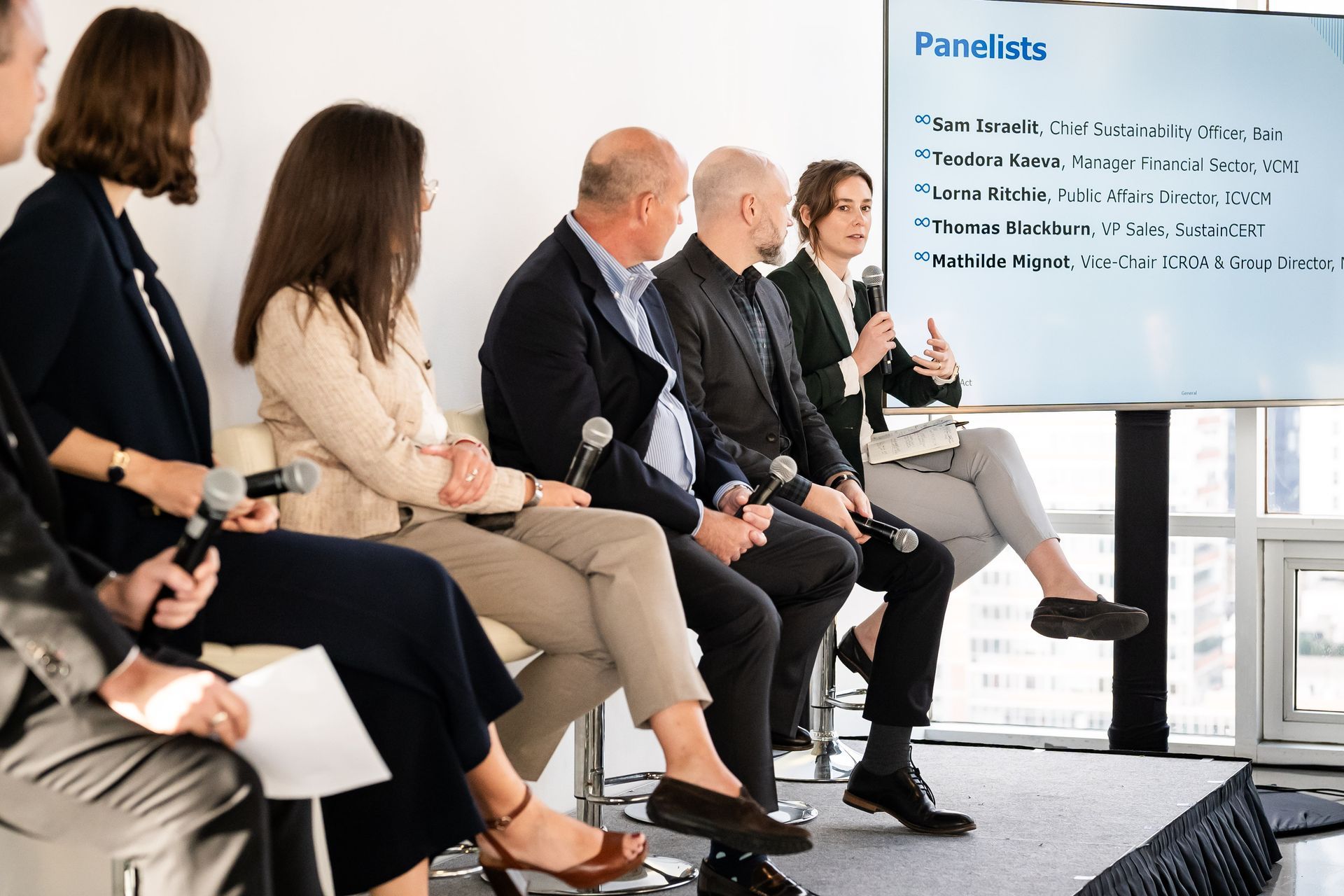 Panel discussion with six people seated, microphones in hand, in front of a screen displaying a list of panelists.