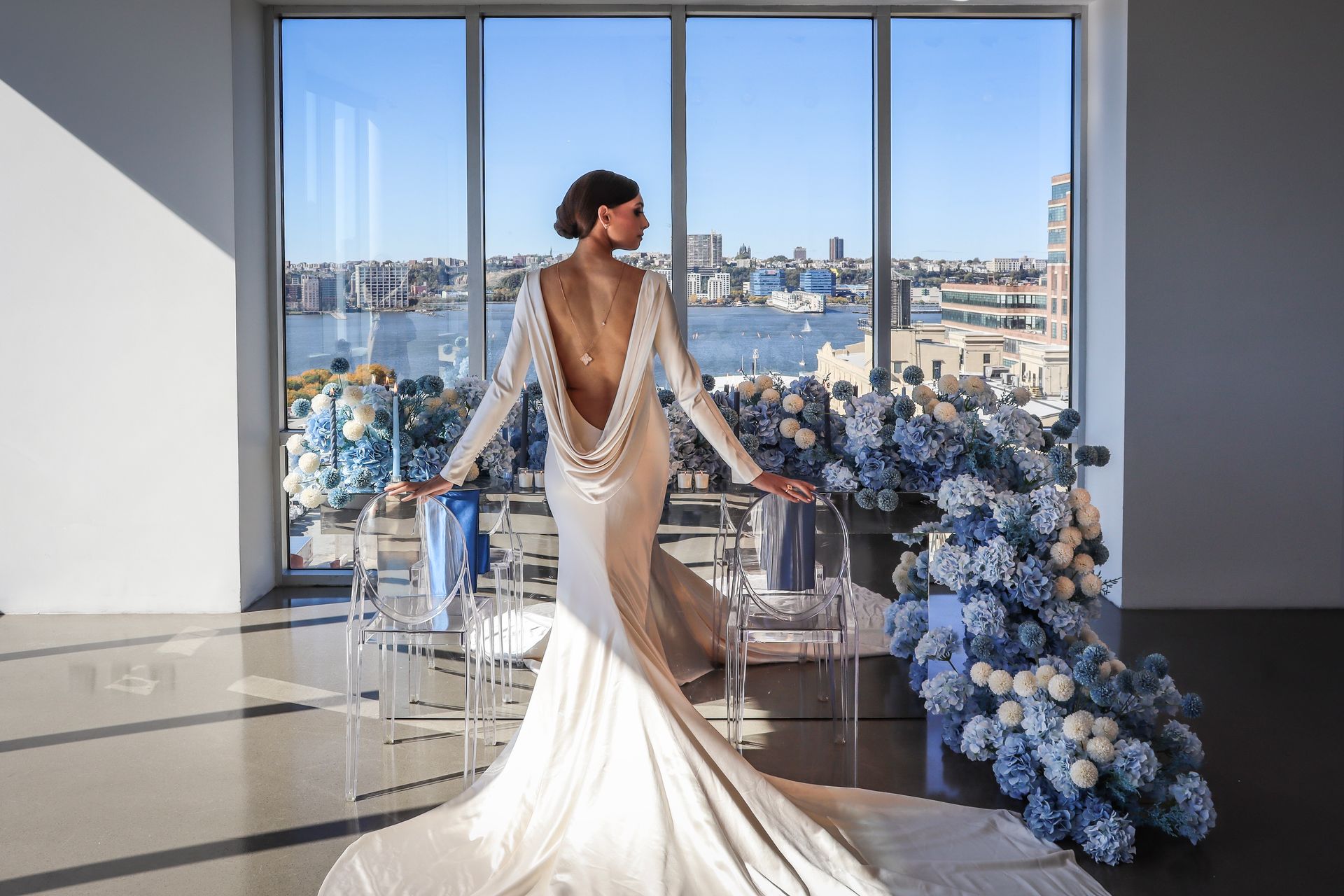 Woman in wedding dress poses by a window overlooking a city and water; blue floral arrangement.
