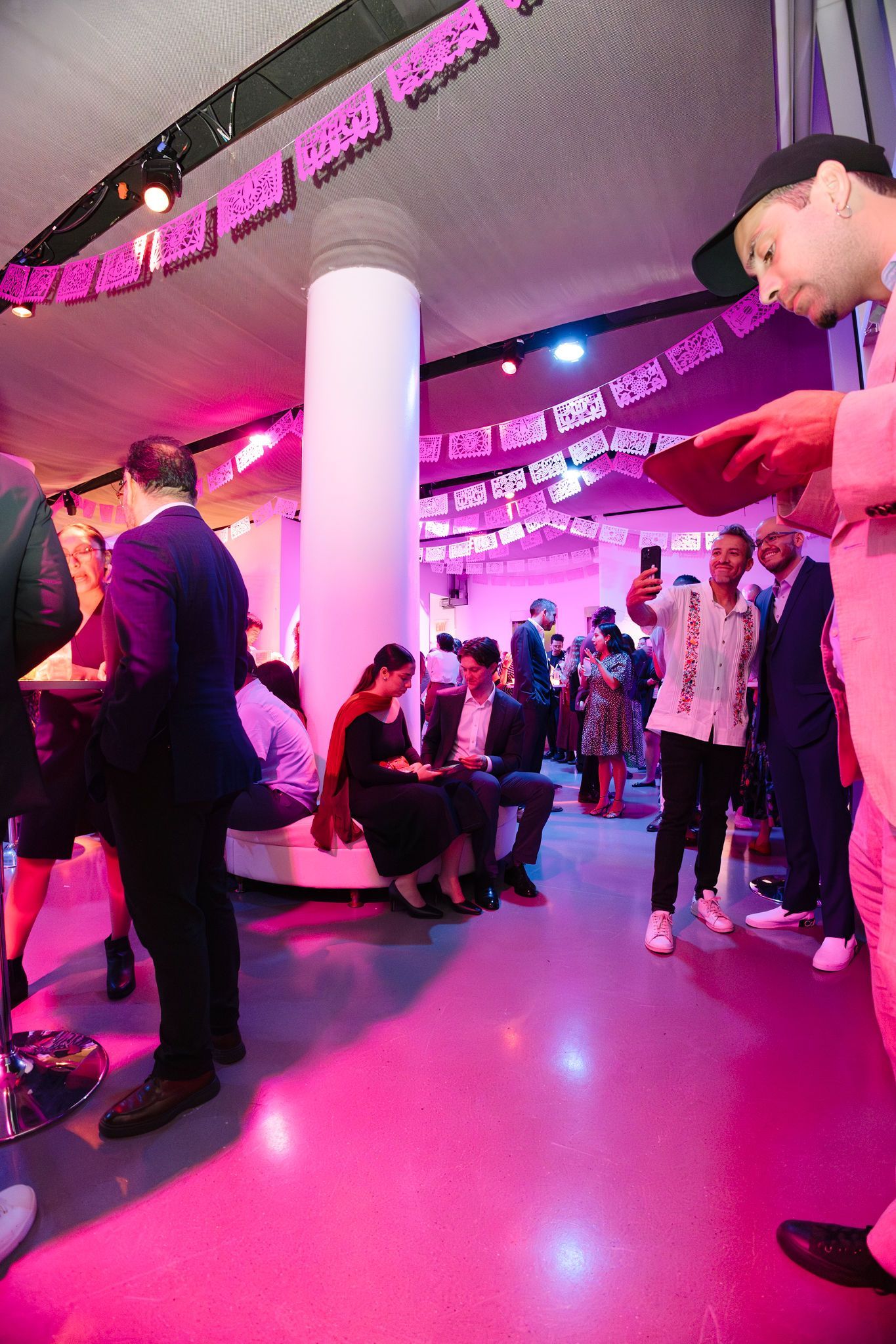 People at an event with pink lighting. A man in a pink suit holds a device. Others converse and take photos.