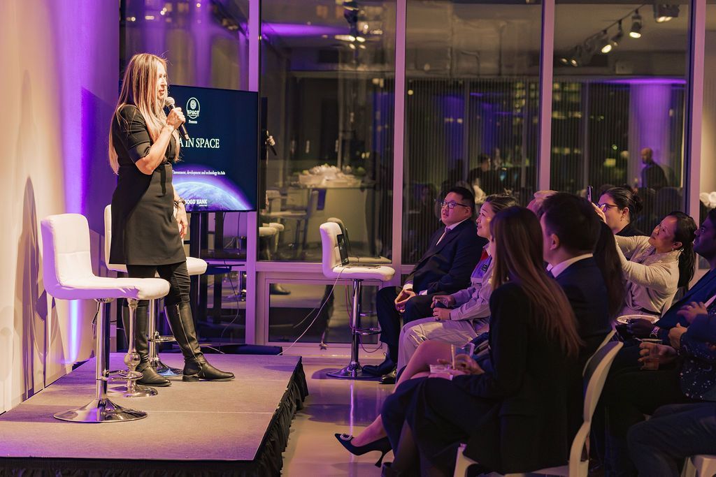 Woman speaking on stage at a venue. Audience seated, looking at speaker. Background has cityscape visible through windows.