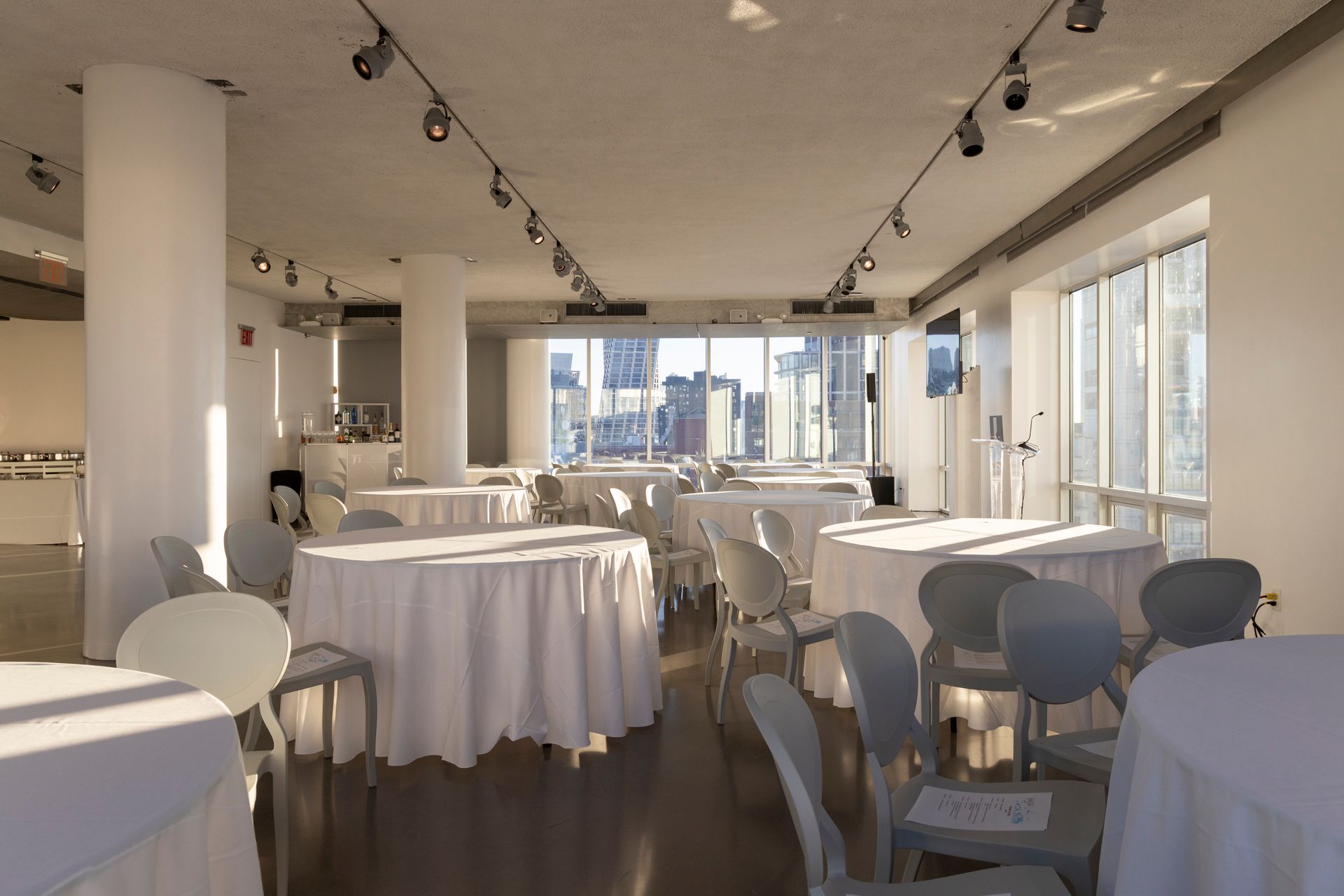 A bright room with round tables covered in white linens and chairs, looking out over a city.
