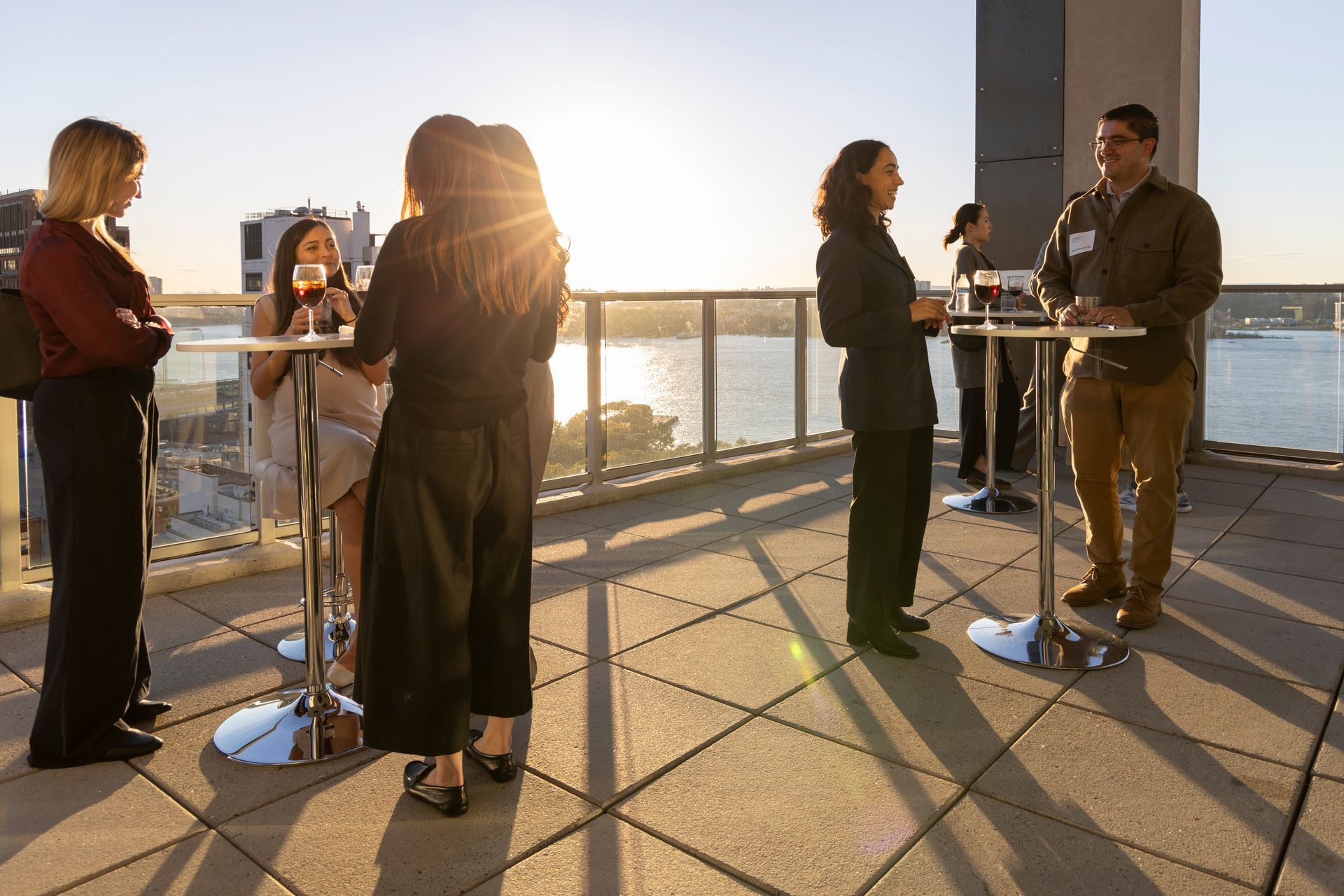 People socializing on a rooftop, enjoying drinks, with a cityscape and water in the background at sunset.