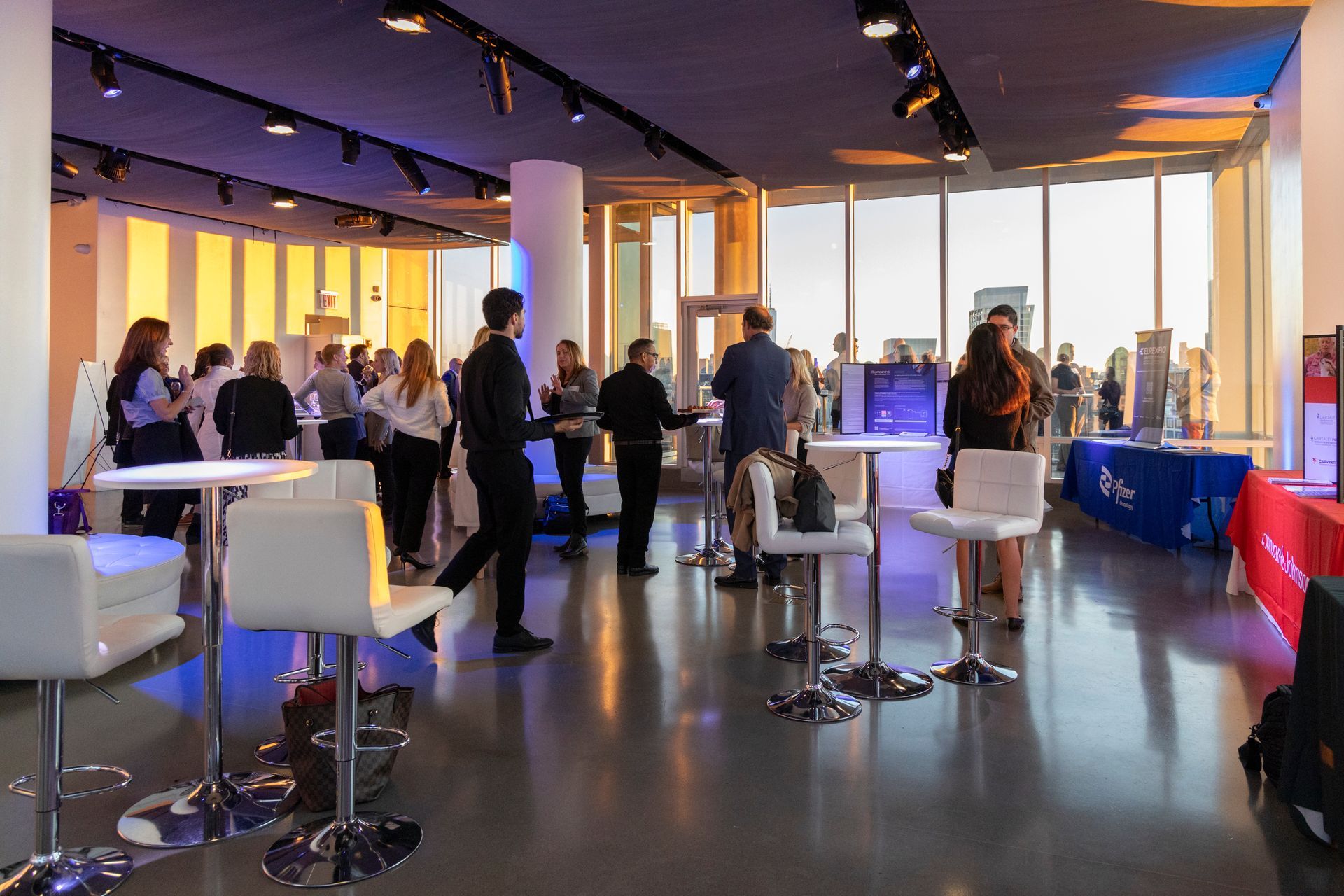 Event with people networking, standing at tables, large windows with city view, and blue and white lighting.