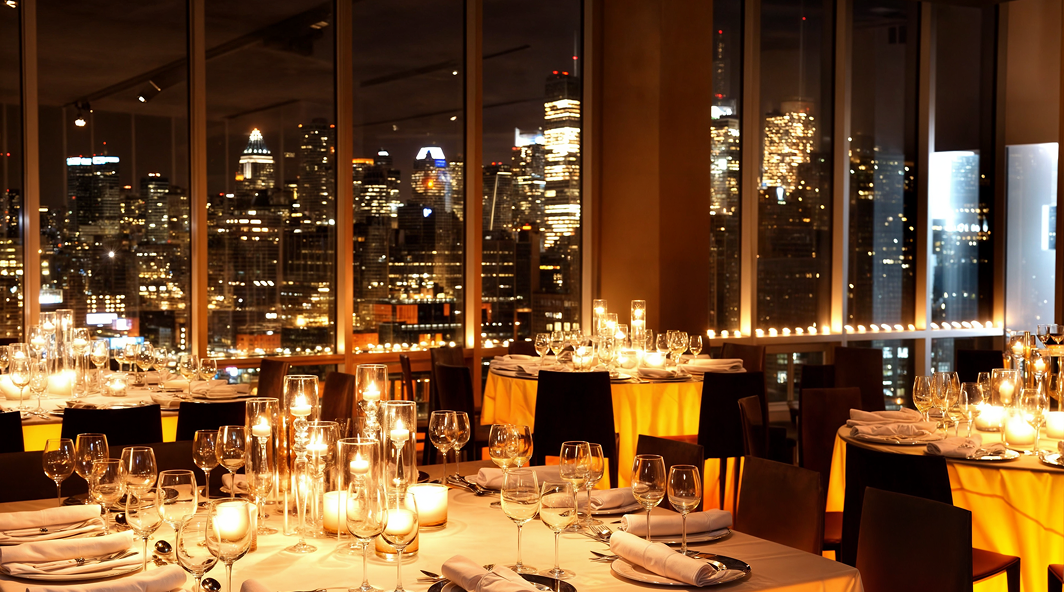 Restaurant interior with tables set for dinner, overlooking a lit-up cityscape at night.