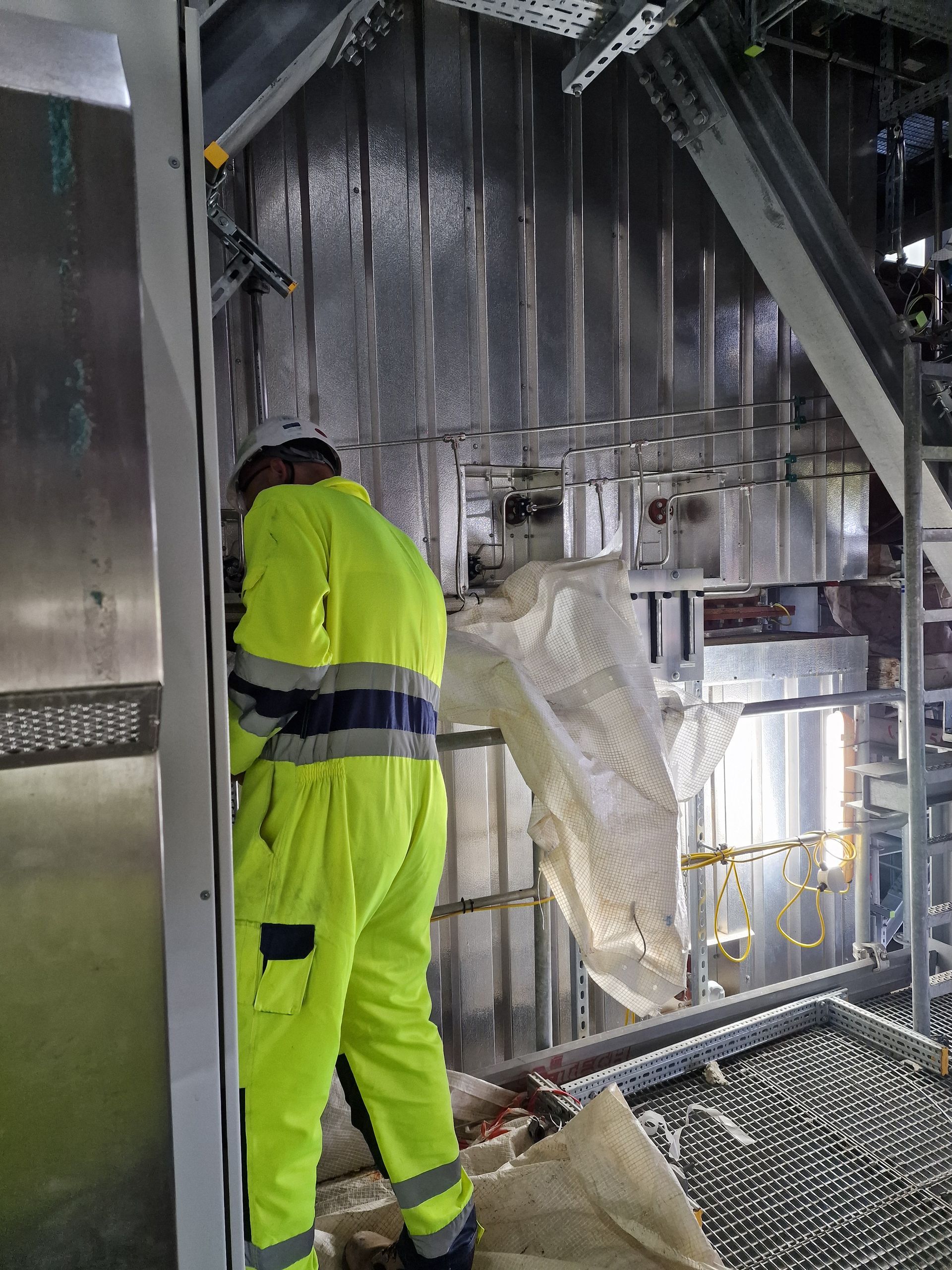 A person in a high-visibility yellow uniform works near metallic industrial equipment in an indoor facility.