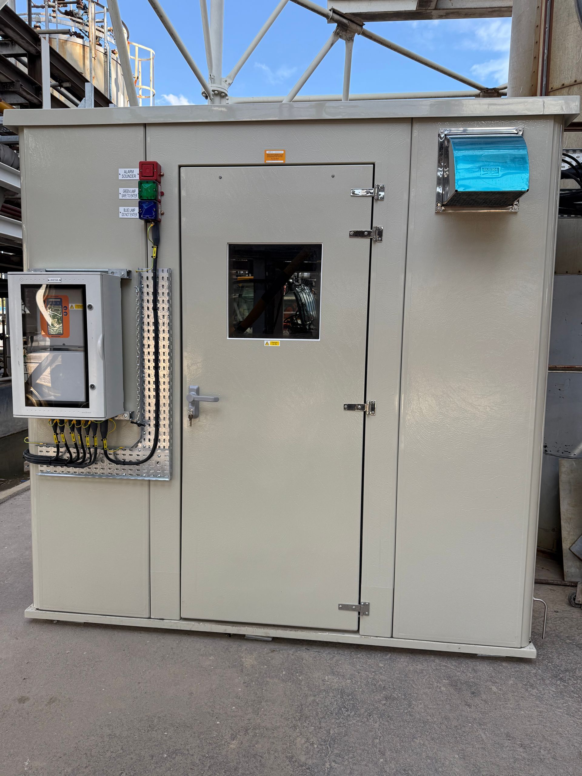 A beige, industrial equipment enclosure featuring a central door with a window, a side control panel, and indicator lights.