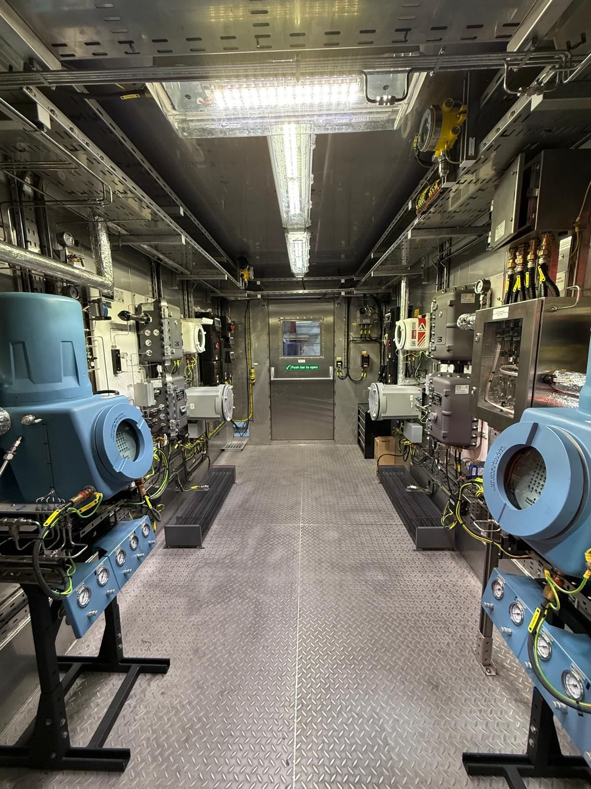 An industrial equipment room with blue machinery on stands lining the sides, a metal floor, and a door at the back.