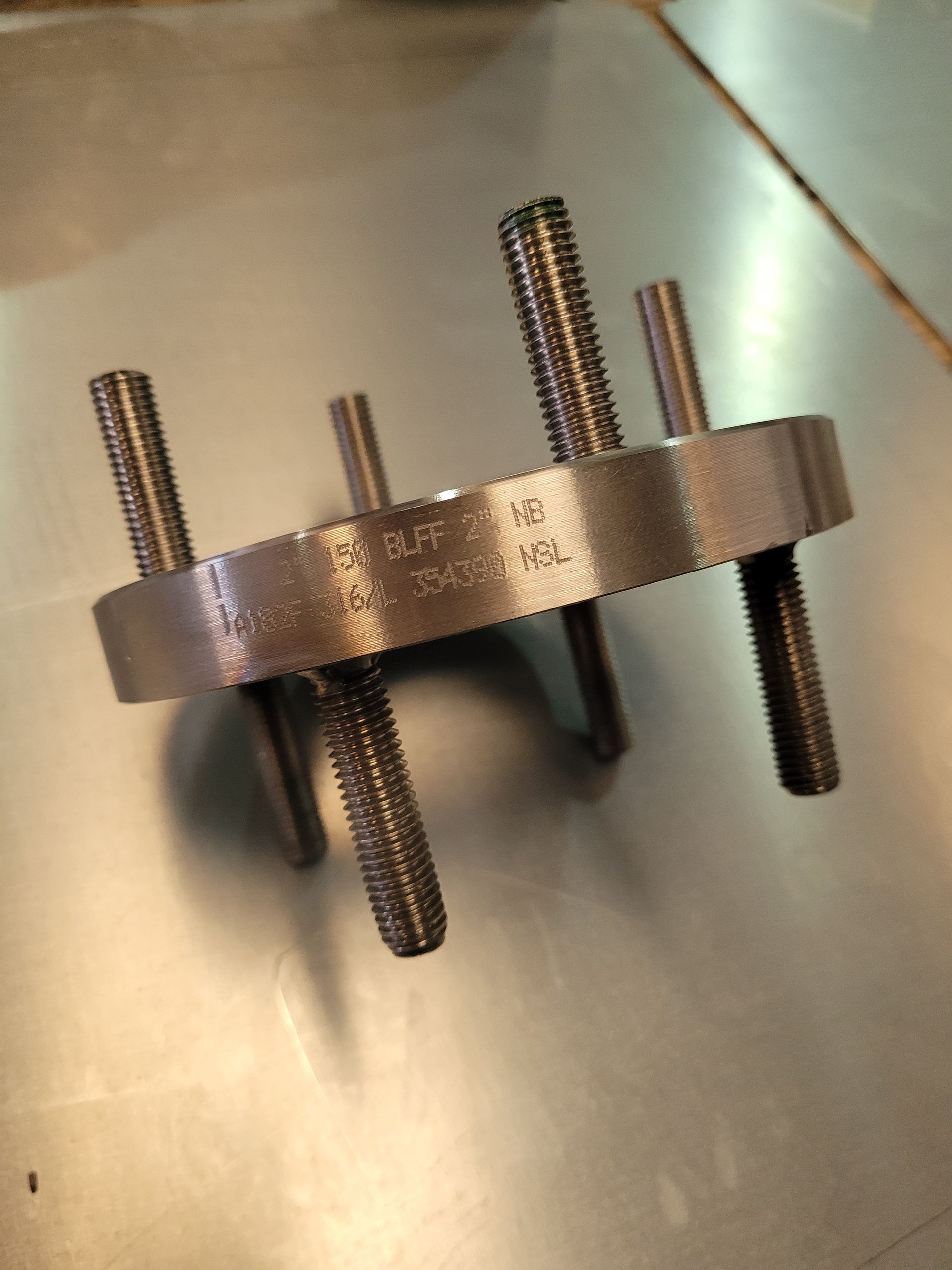 A metal circular flange with threaded studs protruding from one side, resting on a flat, reflective surface.