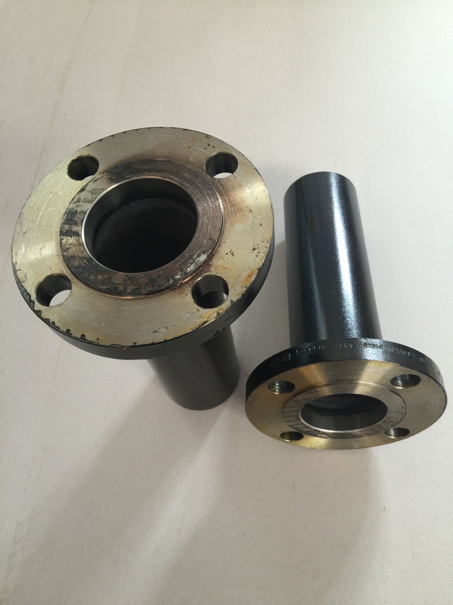 Two black, flanged metal pipe fittings sitting on a white surface, one upright and one angled.