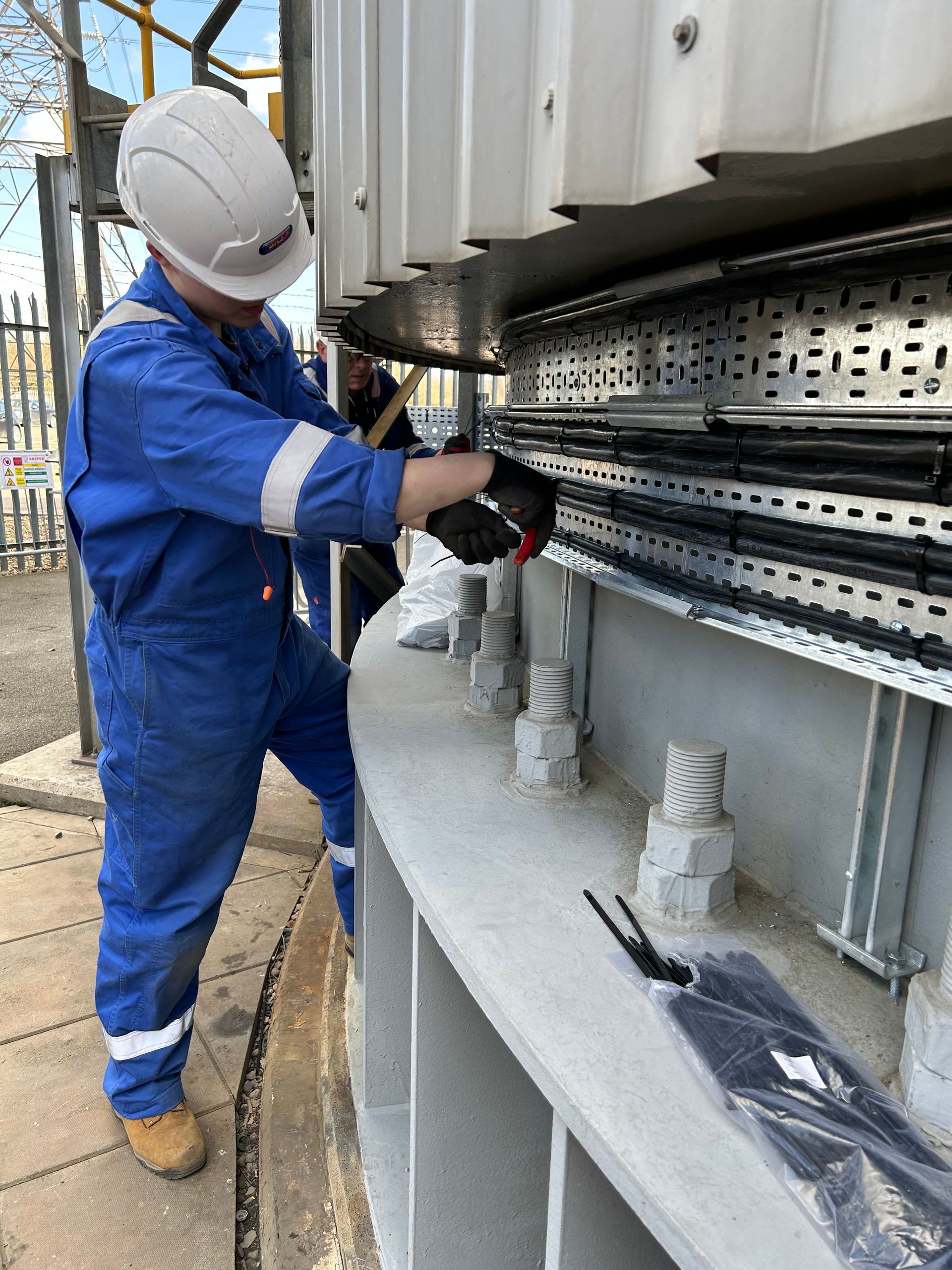 A worker in a blue jumpsuit and hard hat installs or repairs electrical cables in a tray along a large, curved structure.