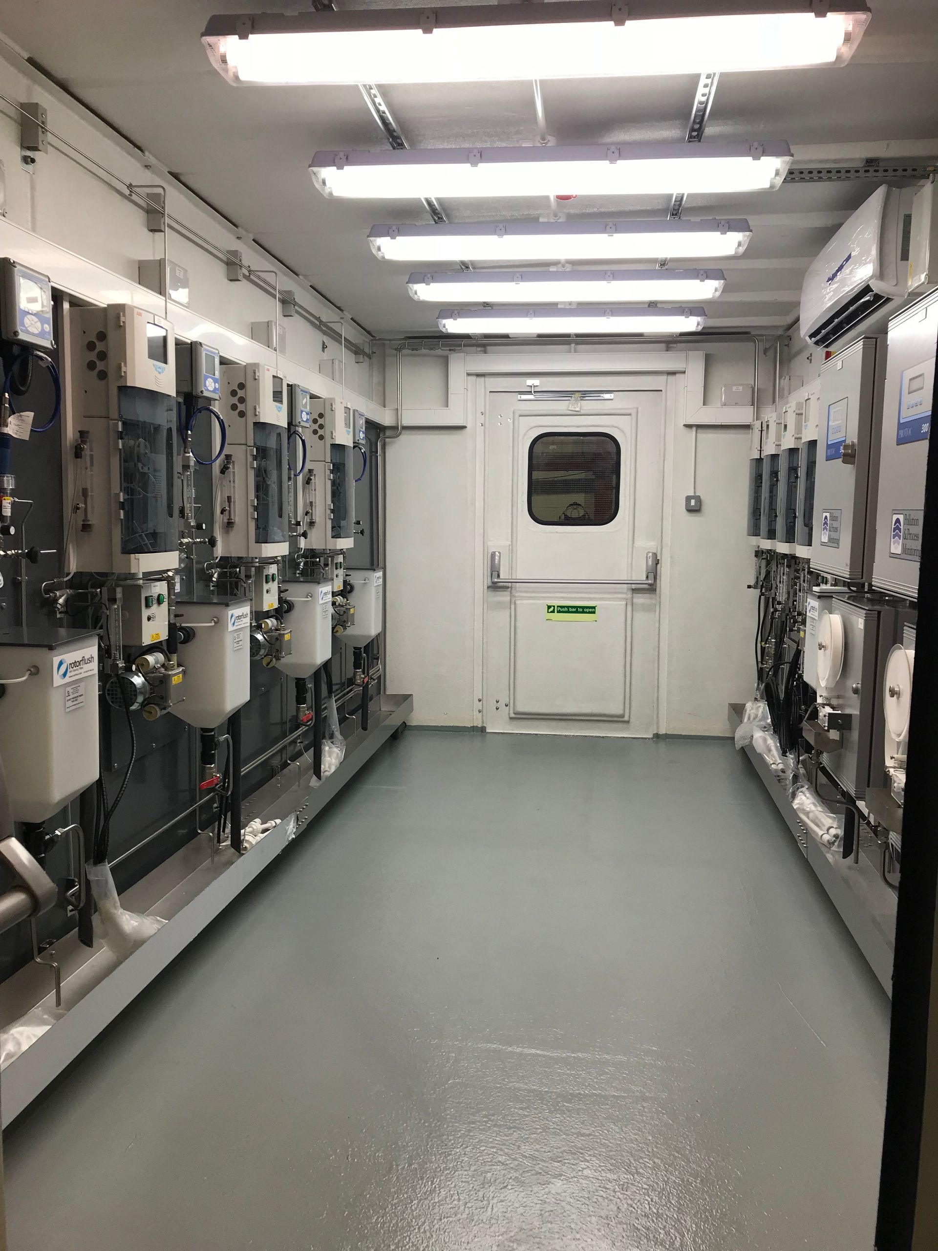 Narrow hallway lined with industrial electrical equipment on gray walls, bright overhead lighting, and a white exit door.
