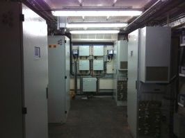A narrow utility room with rows of gray metal electrical cabinets lining the walls and bright fluorescent lighting overhead.