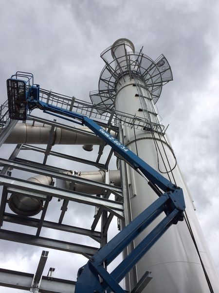 A blue boom lift extends toward a tall industrial exhaust stack against a cloudy sky.