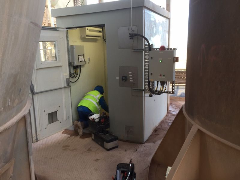 A worker in a high-visibility vest kneels to perform maintenance inside a gray utility shed at an industrial site.