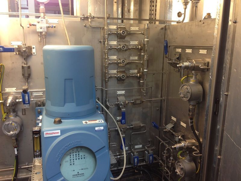 Industrial control panel featuring a blue instrument housing, metallic plumbing, valves, and gauges on a metal wall.