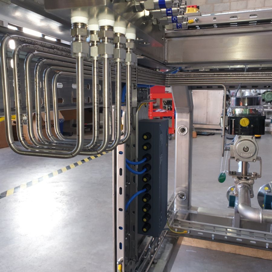 Stainless steel piping loops connected to a manifold above a grey electrical control box in an industrial workshop.