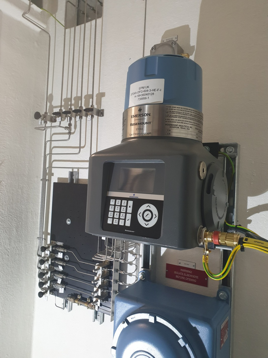 A wall-mounted industrial flow meter or pressure transmitter with a digital display and keypad, connected to metal tubing.