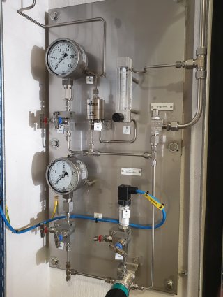 A stainless steel industrial instrument panel with pressure gauges, valves, tubing, and a flow meter mounted on a wall.