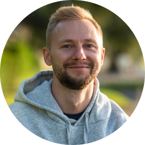 A person with short blond hair and a trimmed beard wearing a grey hoodie, smiling against a soft, sunlit outdoor background.