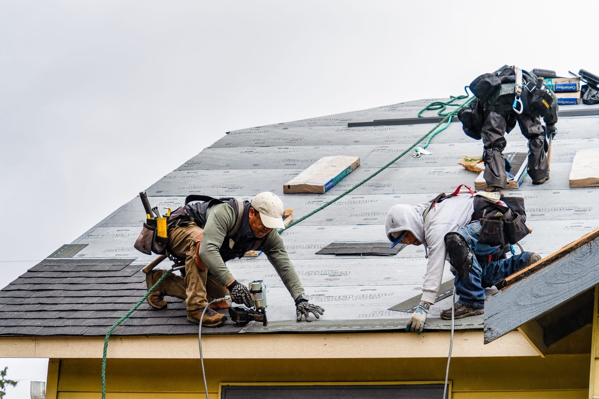 Workers installing roofing on a steep roof, using safety harnesses and ropes.