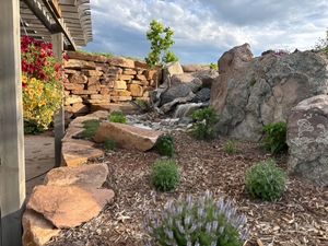 A landscaped backyard features a stone waterfall, tiered rock retaining walls, lush green plants, and a wooden pergola.
