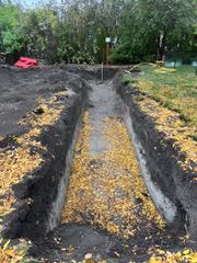 A long, narrow trench excavated into the dirt of a backyard, scattered with yellow autumn leaves.