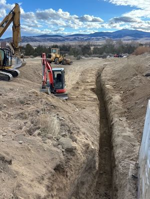 A red excavator digs a trench at a construction site with yellow heavy machinery and distant mountains in the background.
