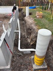 Construction site showing a concrete foundation wall, white PVC pipes, and a cylindrical concrete pillar with a yellow base.