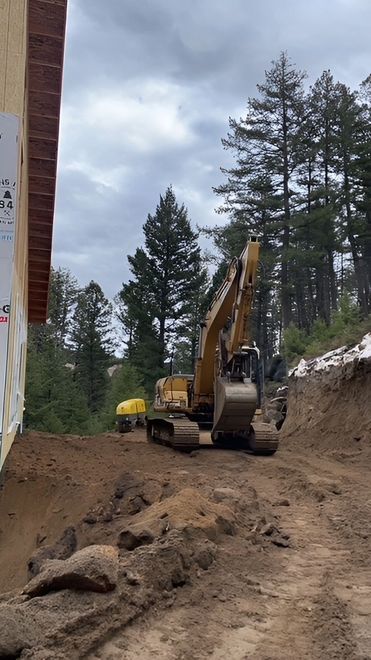 A yellow excavator performs excavation work in a dirt lot beside a building under construction near a forest.