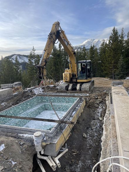 An excavator lifts a tiled pool shell from an excavated site in a snowy, mountainous, pine-forested landscape.