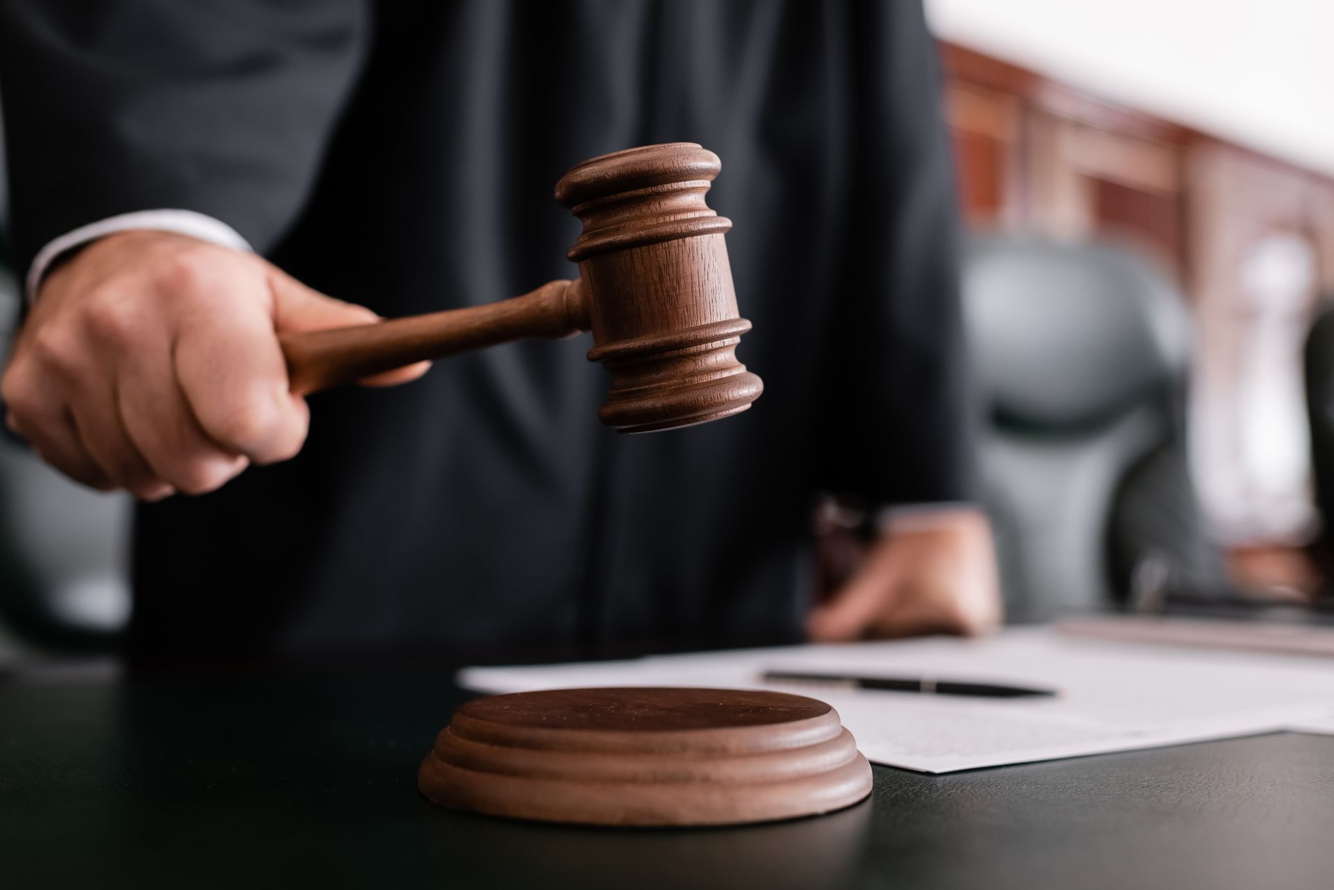 A judge in a black robe holding a wooden gavel over a sounding block on a courtroom desk.