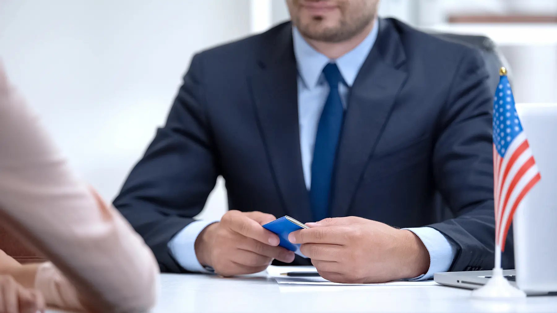 Man in suit holding a blue card, seated across from someone at a table, American flag in background.