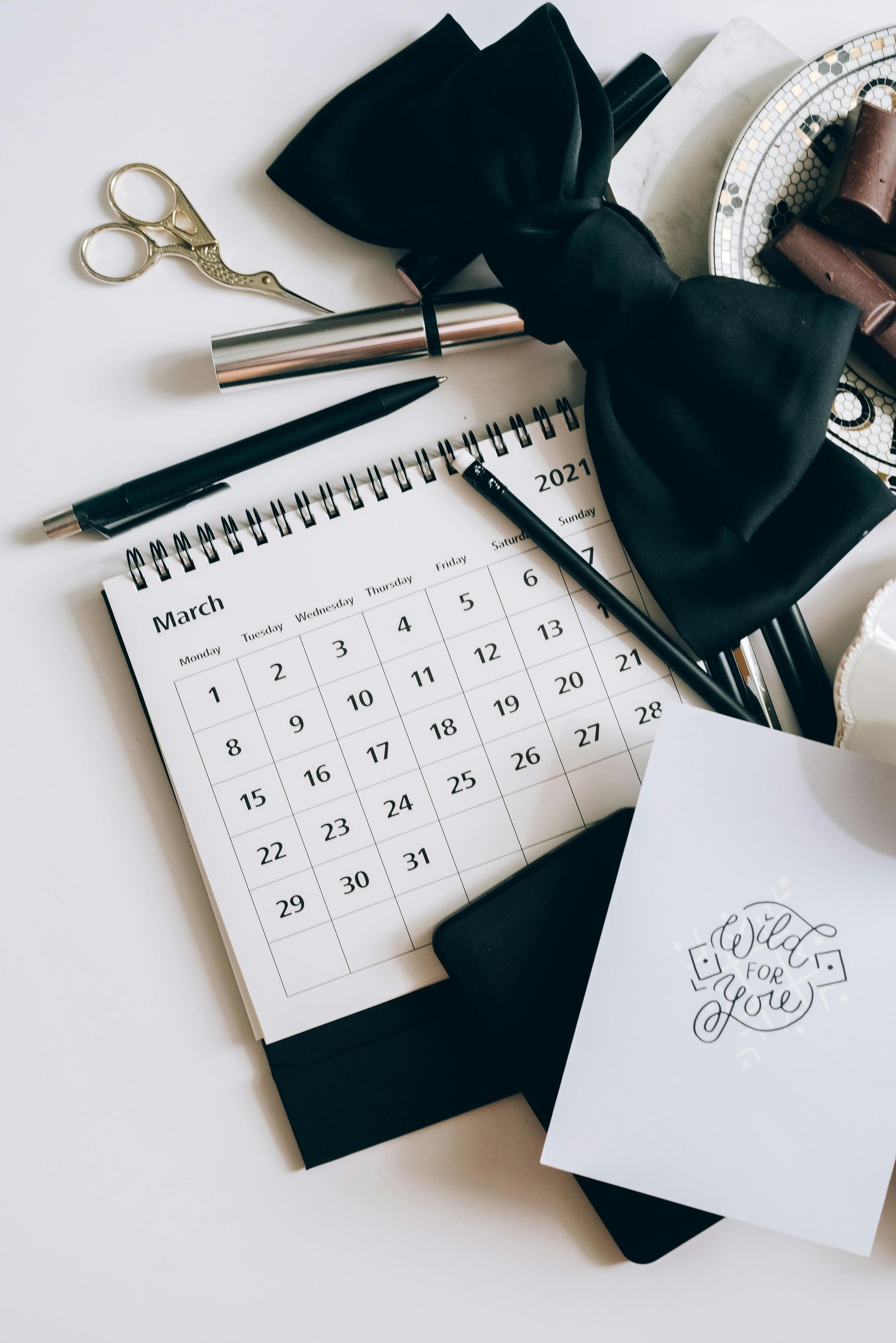 A calendar , pen , scissors , and a bow tie are on a table.
