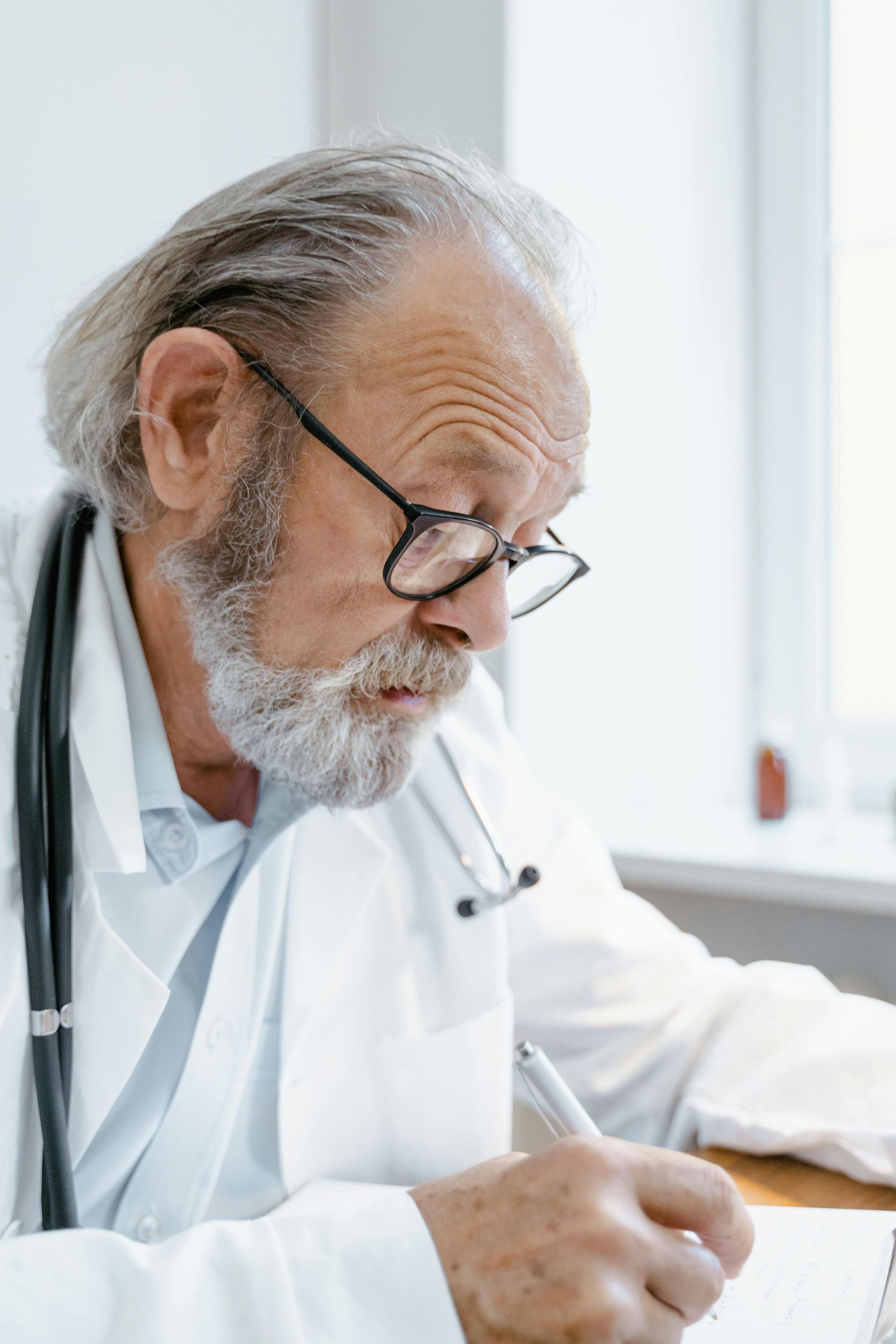 An elderly doctor is sitting at a desk writing in a notebook.
