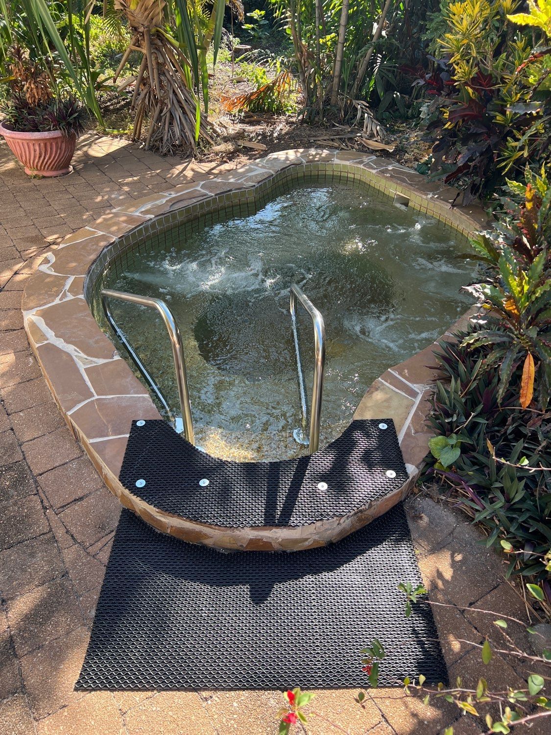 Small outdoor spa with bubbling water, surrounded by stone and greenery. Chrome handrails and a black mat  — Friendly Pool & Spa Care in Larrakeyah, NT 