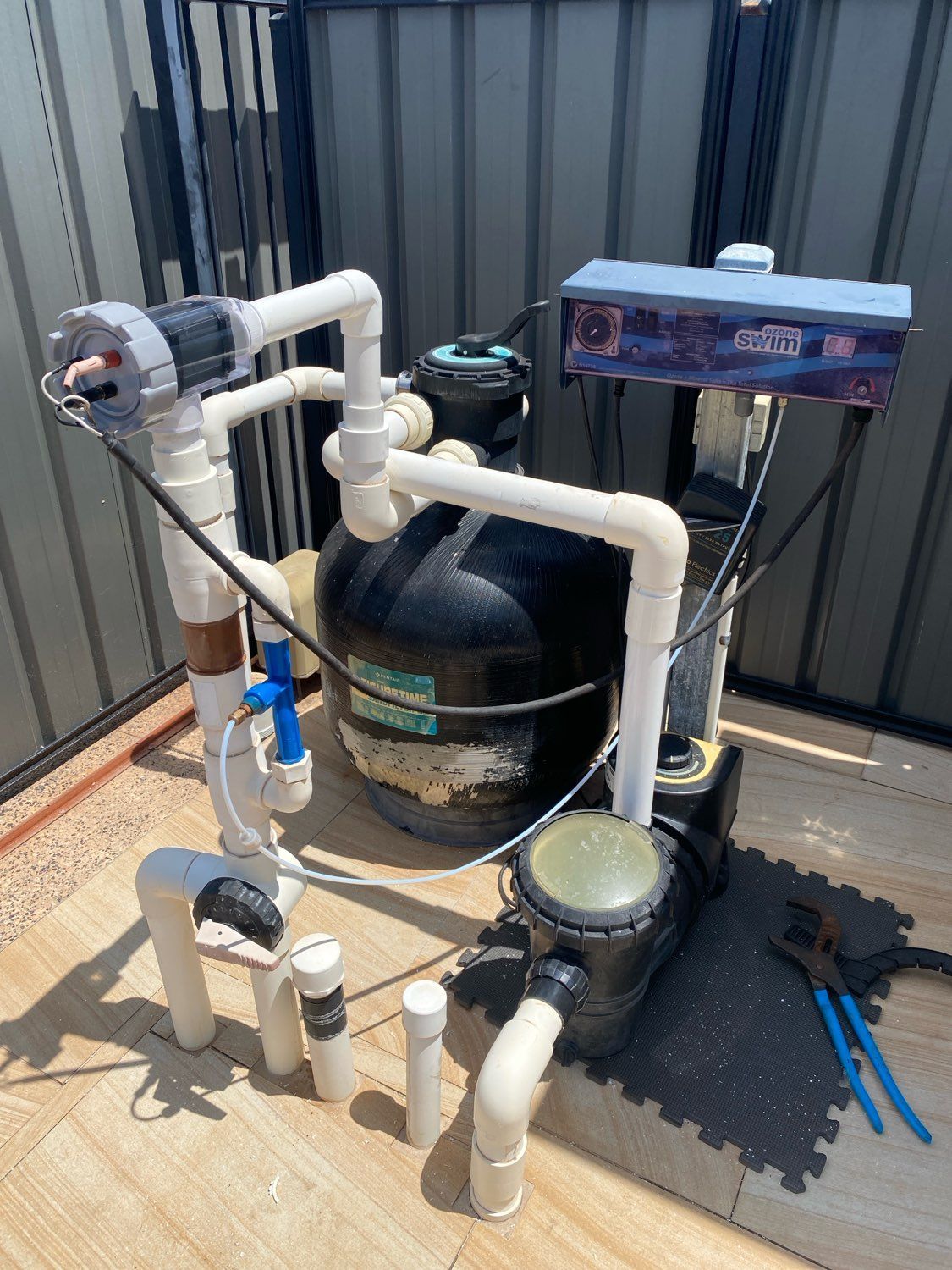 Swimming pool filtration system with pump, black tank, and saltwater chlorinator against a grey fence on a tiled patio   — Friendly Pool & Spa Care in Yarrawonga, NT 