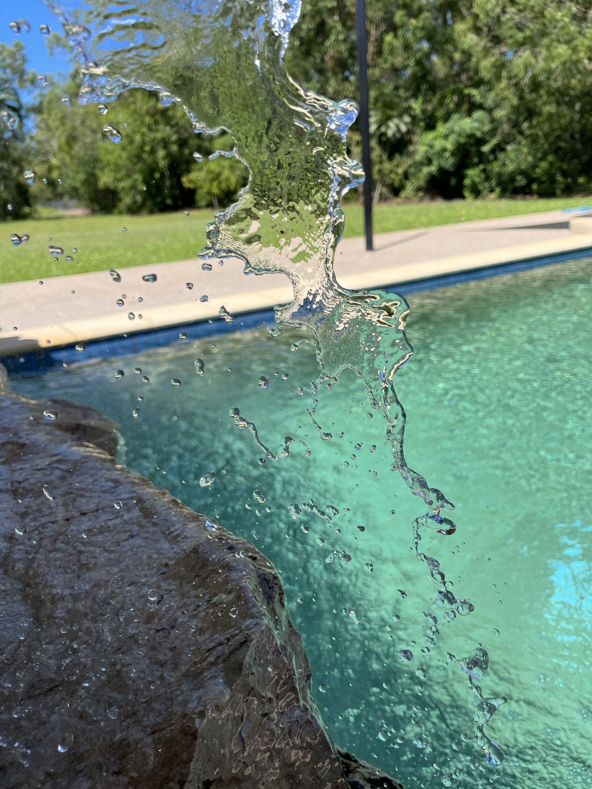Water cascades from a dark, jagged rock ledge into a turquoise backyard swimming pool under a bright sky  — Friendly Pool & Spa Care in Coconut Grove, NT 