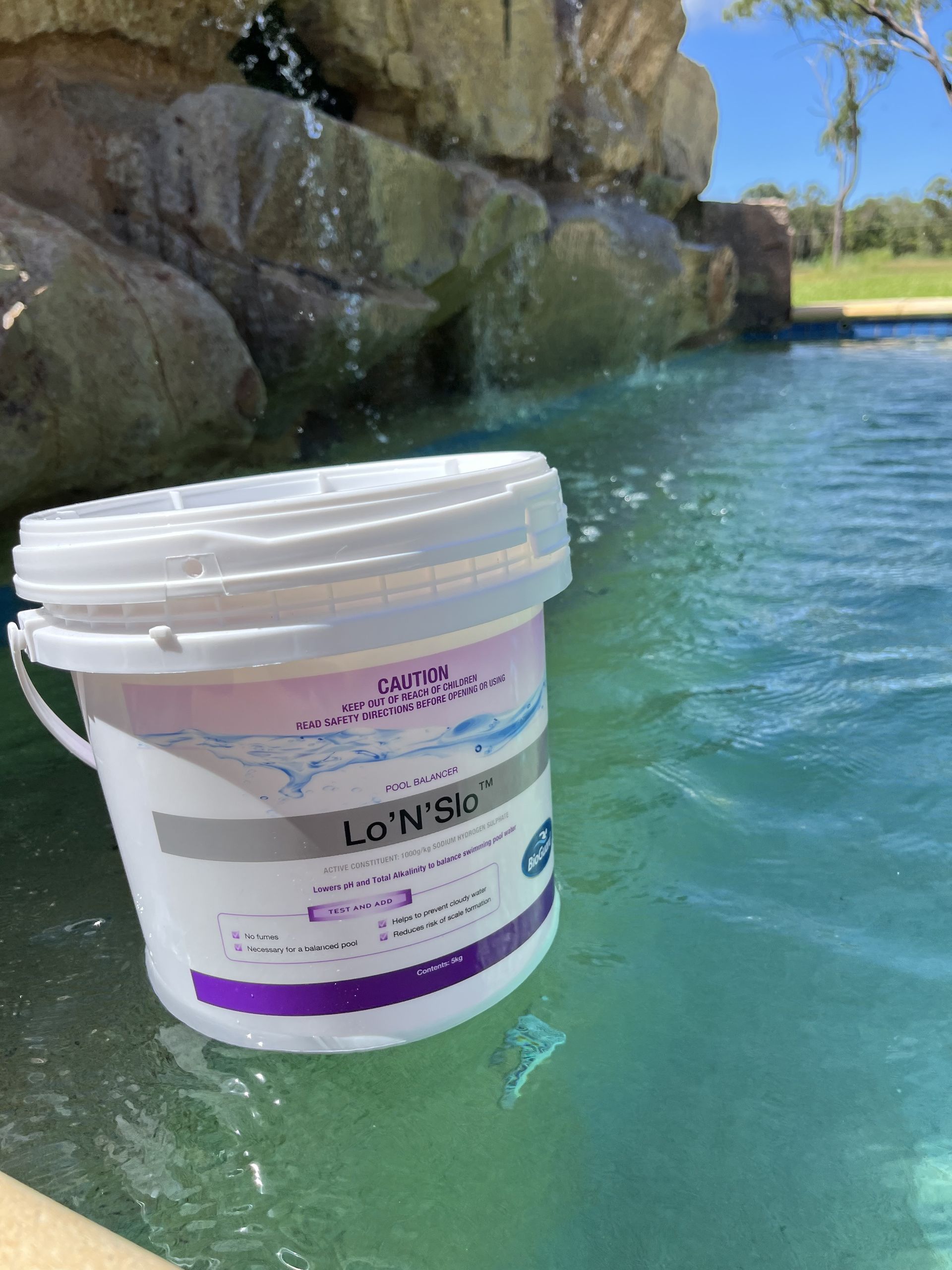 A white bucket labeled Lo-N-Slo sits on the edge of a backyard swimming pool with a stone waterfall feature — Friendly Pool & Spa Care in Coconut Grove, NT 
